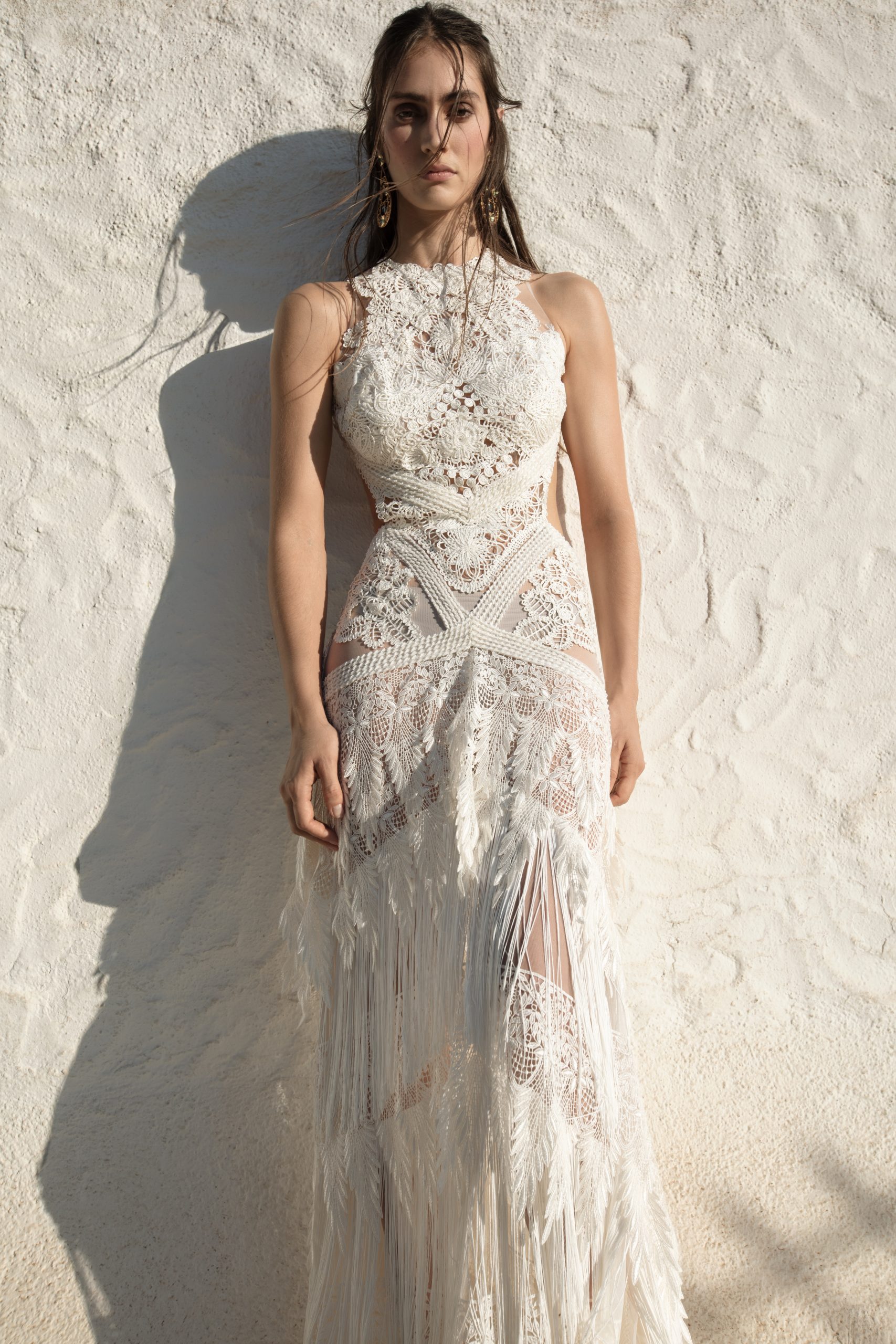Modelo posa para foto com vestido de noiva boho, na cor branca, de corpete com alças de renda, atrás dela um muro chapiscado.