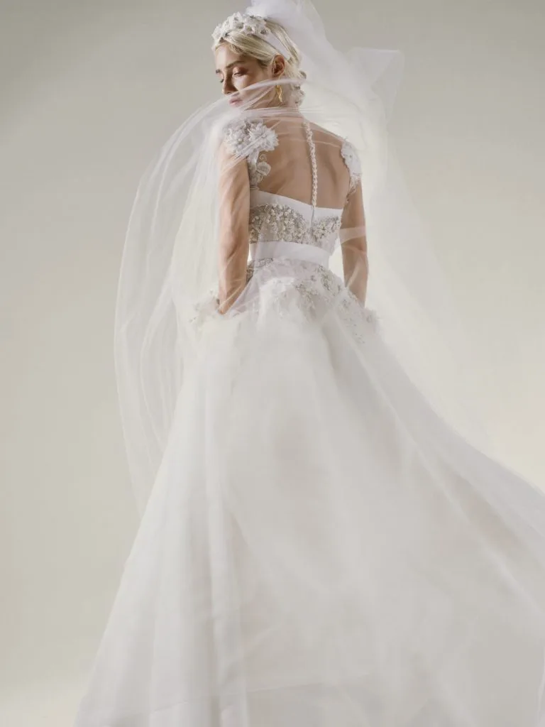 Mulher usando um vestido de noiva branco com detalhes floridos nas mangas e uma tiara de flores na cabeça de onde sai um lindo véu.