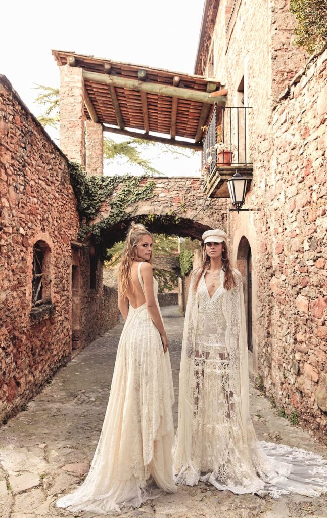 Duas mulheres em pé, posando em vestidos de noiva rendados. A modelo à direita veste boina e a segunda está inclinada à foto.