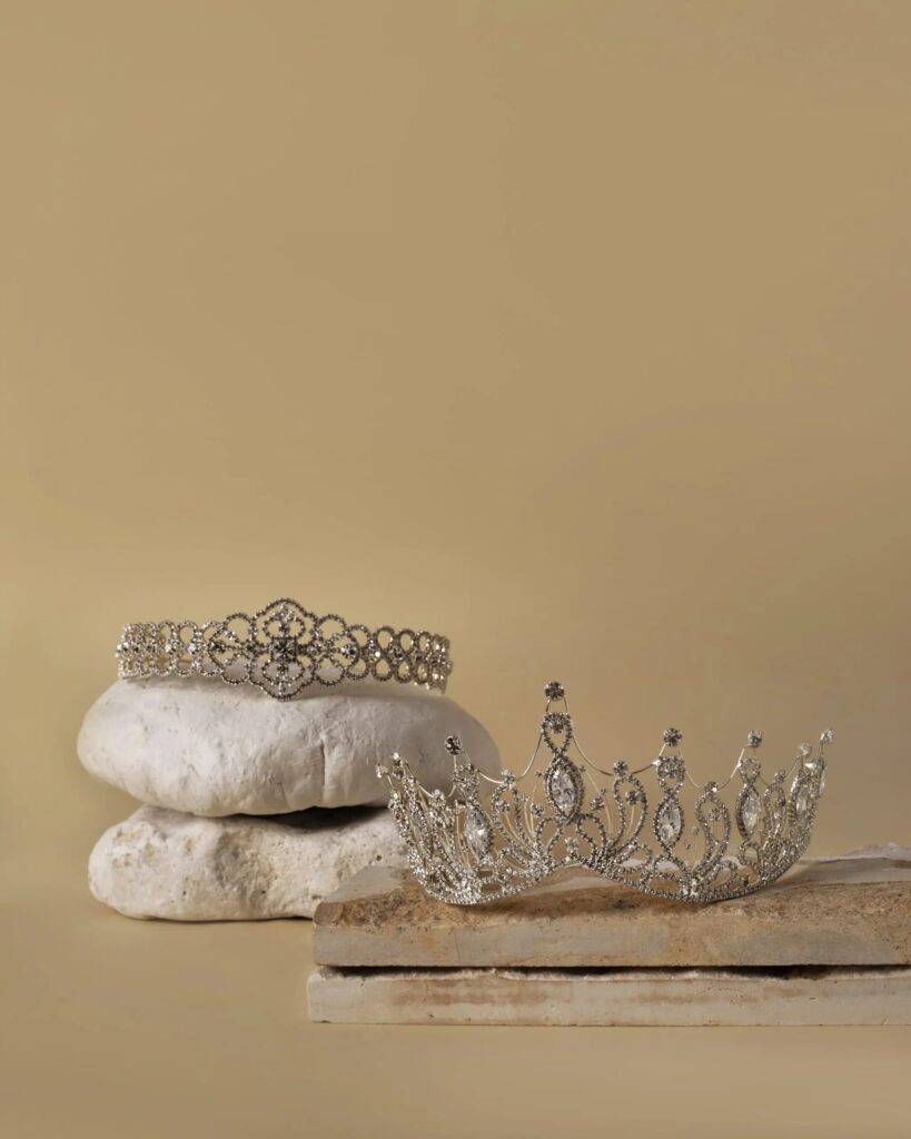 Uma tiara e uma coroa, ambas cravejadas em pedras brilhantes, apoiadas em uma base de pedra bege, em um fundo no mesmo tom.