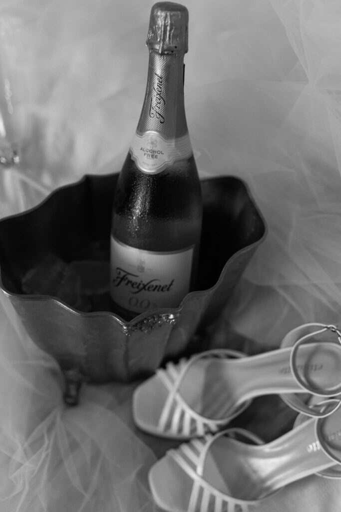 Imagem em preto e branco de um par de sandálias de salto-alto e um balde de gelo com garrafa de champagne dentro.