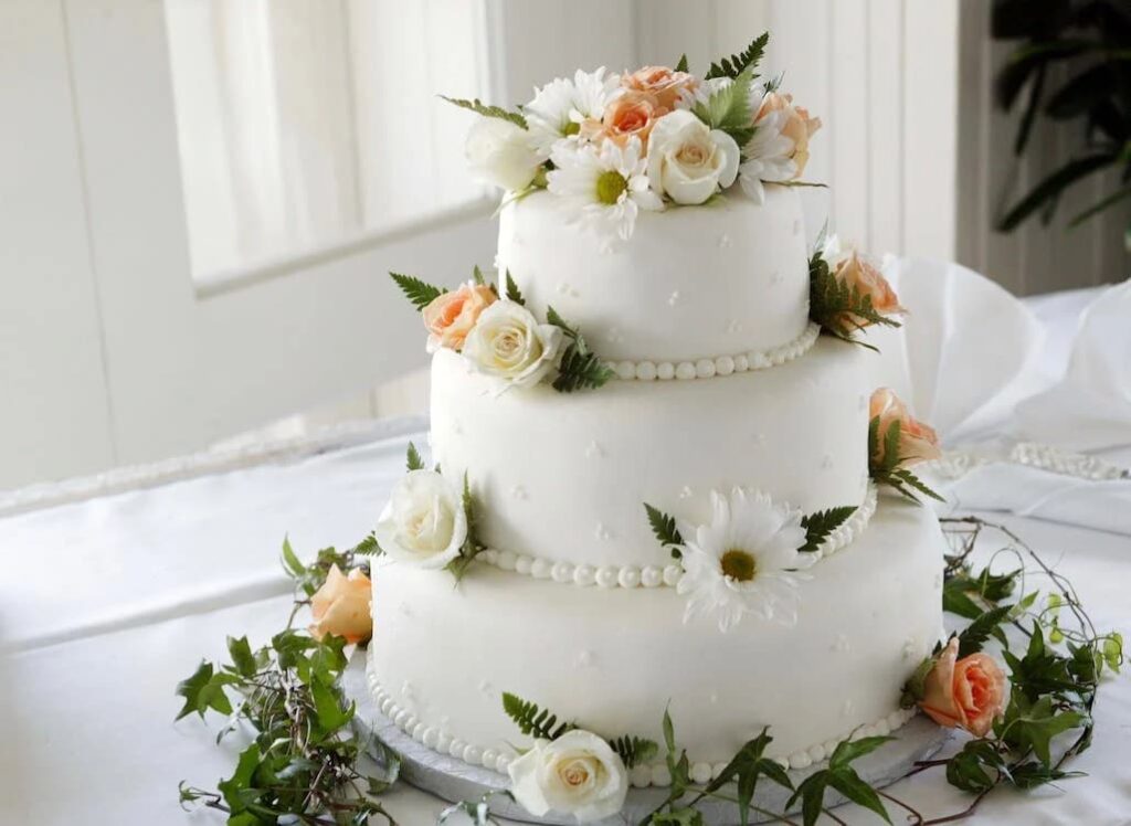 Bolo de casamento clássico de três andares, com cobertura branca e decoração feita por flores brancas, rosas e folhas verdes.