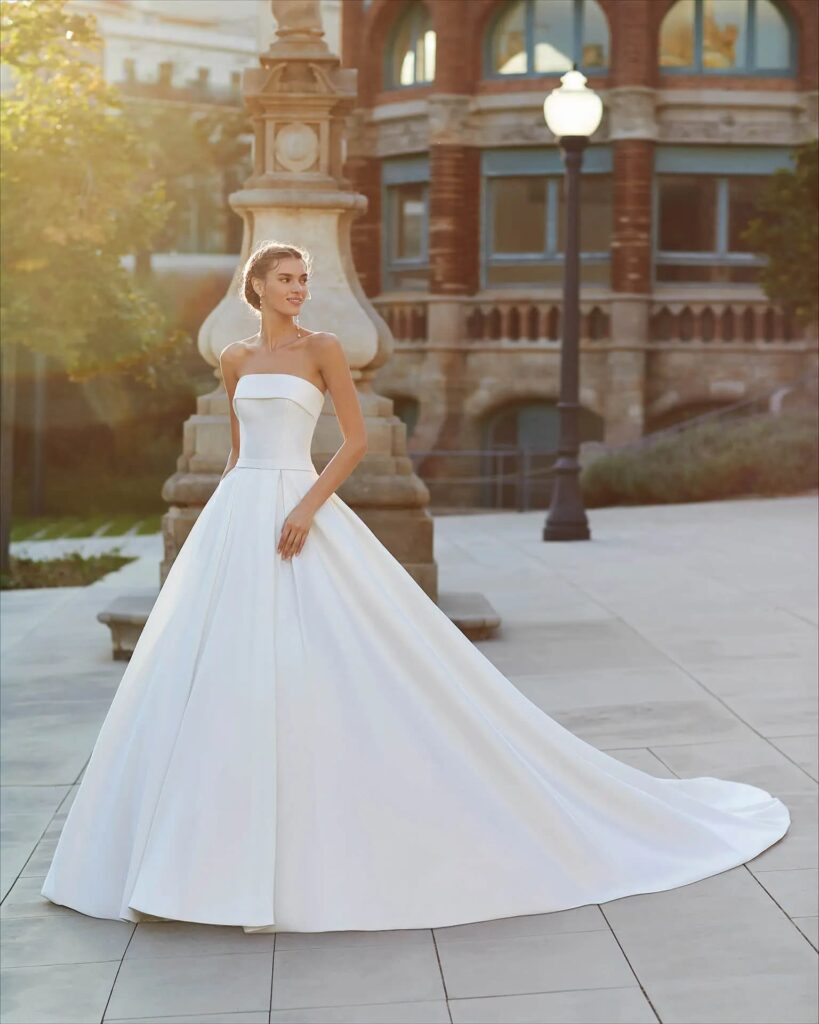 Modelo usando vestido de noiva clássico, branco, tomara que caia e com tecido liso, posando em praça pública, à luz do sol.