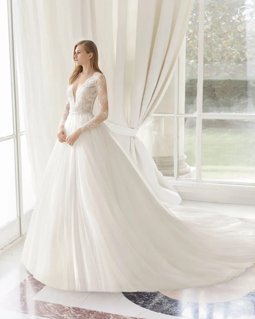 Modelo usando vestido de noiva clássico, branco, com mangas longas e tecido rendado, em frente a janela com cortinas brancas.