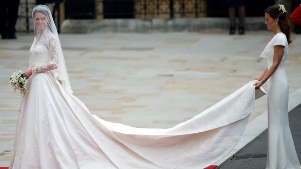 Kate Middleston em seu vestido de noiva, sorrindo para a câmera enquanto uma mulher segura a cauda do vestido, logo atrás.