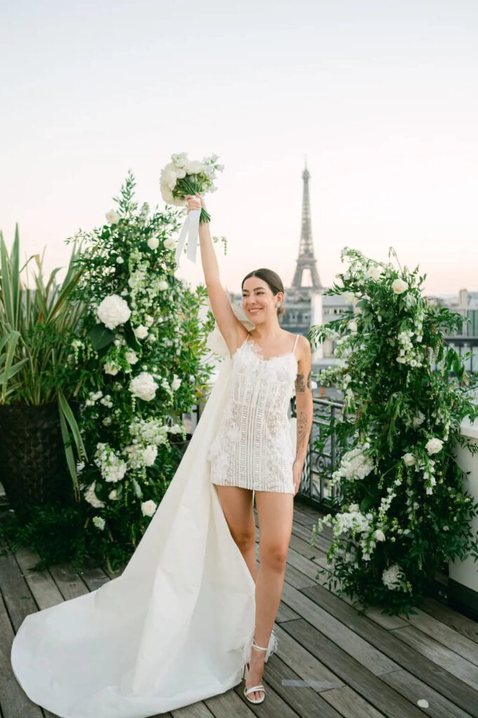 Nath Araújo posam para foto em frente à Torre Eiffel, utilizando vestido de noiva Yolancris curto feito em renda e guipura, com aplicações florais e com uma cauda caindo do ombro com um laço largo.
